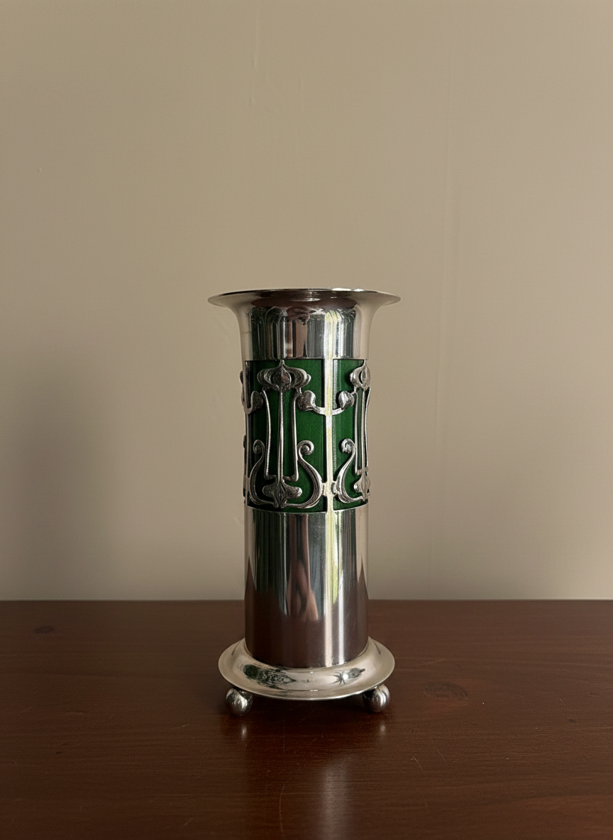 A polished silver-plated pedestal vase with a decorative art nouveau green insert and cutout scrollwork, standing on a dark wooden table against a plain beige wall.