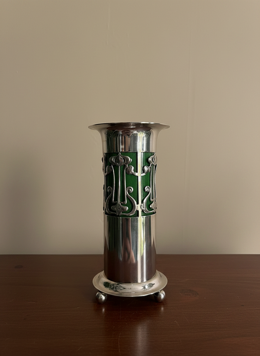 A polished silver-plated pedestal vase with a decorative art nouveau green insert and cutout scrollwork, standing on a dark wooden table against a plain beige wall.