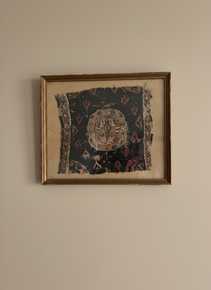 a framed piece of coptile textile featuring two figures mounted on a brown backing on a taupe coloured wall