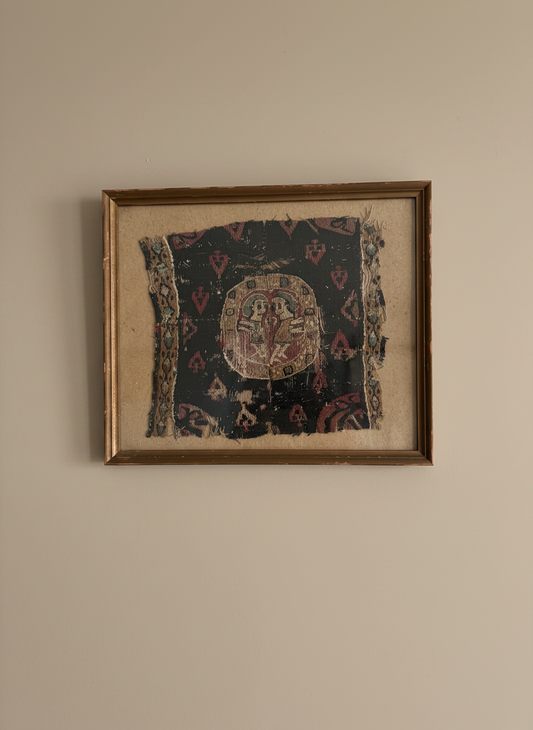 a framed piece of coptile textile featuring two figures mounted on a brown backing on a taupe coloured wall