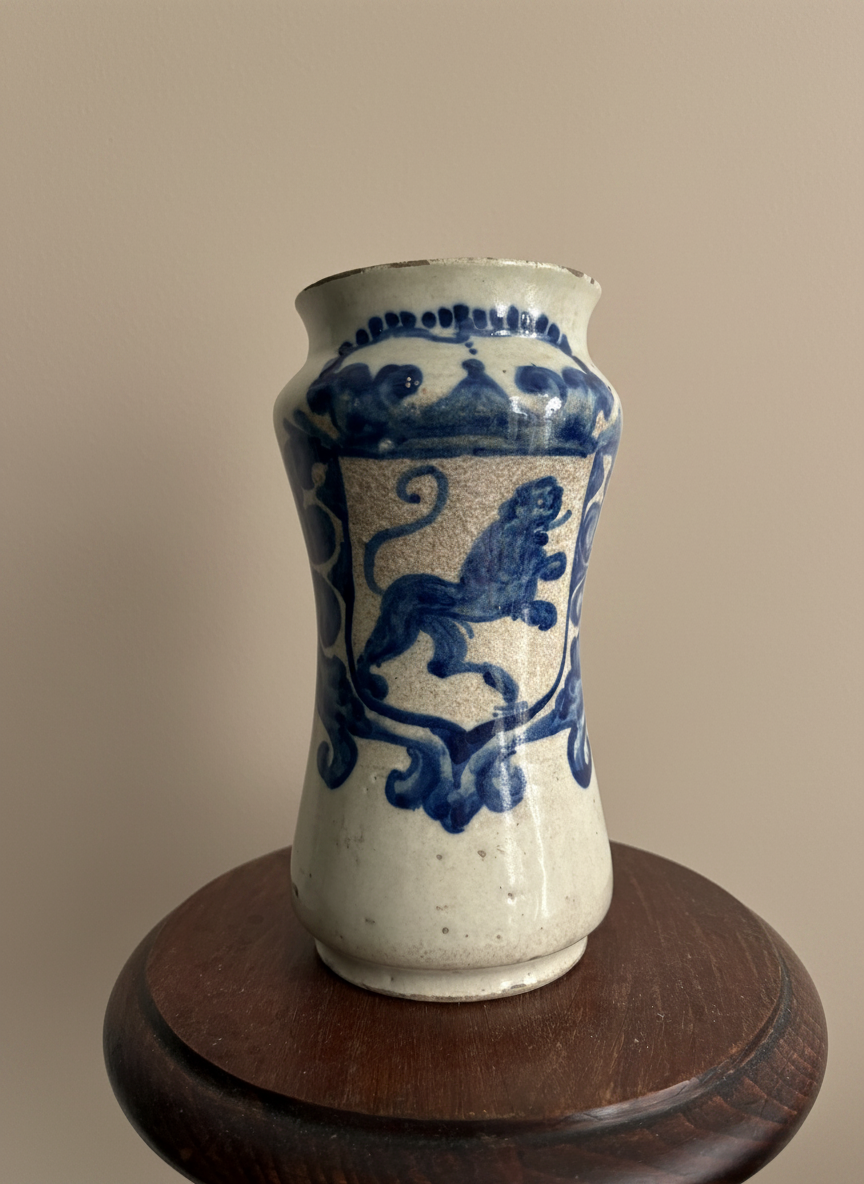 close up of a maiolica vase in off white with a blue rampant lion on a shield. placed on a brown wood table against a taupe wall